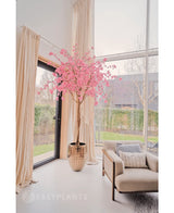 L'arbre rose en fleurs de cerisier artificiel 240 cm se dresse dans un pot décoratif près d'une grande fenêtre dans un salon lumineux et moderne avec des rideaux couleur crème, un fauteuil beige et une vue sur le jardin extérieur.