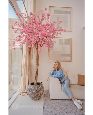 Une femme vêtue de vêtements décontractés se détend sur un canapé beige à côté d'un cerisier rose artificiel de 240 cm avec des fleurs roses, placé dans un salon moderne avec des œuvres d'art murales abstraites et de grandes fenêtres.