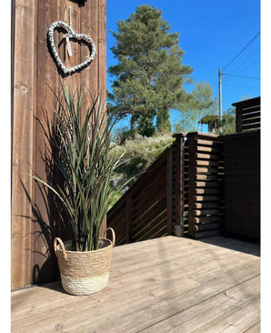 Le gazon artificiel Bordeaux UV 120 cm est posé dans un panier tressé sur une terrasse en bois près d'un mur en bois avec une couronne en forme de cœur, avec des arbres et un ciel bleu clair en arrière-plan.