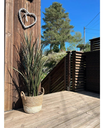 Le gazon artificiel Bordeaux UV 120 cm est posé dans un panier tressé sur une terrasse en bois près d'un mur en bois avec une couronne en forme de cœur, avec des arbres et un ciel bleu clair en arrière-plan.