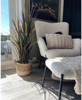 Un coin cosy avec un fauteuil moelleux blanc, un pouf assorti, un coussin texturé et du gazon artificiel Bordeaux UV 120 cm dans un panier tressé avec la lumière du soleil qui brille à travers une grande fenêtre.