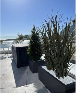 Le gazon artificiel Bordeaux UV 120cm en jardinières rectangulaires avec pierres blanches agrémente une terrasse extérieure moderne sur fond de ciel bleu et d'arbres au loin.