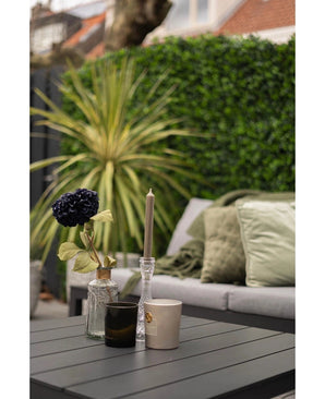 Une table d'extérieur moderne avec une surface noire montre un vase avec une fleur sombre, un bougeoir en verre et deux tasses. Derrière, se trouve un canapé avec des coussins verts, face à un mur végétal luxuriant composé du mélange de tapis Kunsthaag 50x50 cm.