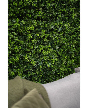 Gros plan d'un tapis de haie artificielle vert luxuriant mélangeant un mur de 50 x 50 cm avec des feuilles vibrantes, avec le coin d'un canapé gris clair et une partie d'un manteau vert visible au premier plan inférieur.