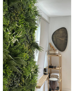 Une haie artificielle luxuriante Fraîcheur de jardin vertical UV 100x100 cm recouvre un mur d'une pièce lumineuse. A proximité, une horloge murale triangulaire moderne est suspendue au-dessus d'une étagère en bois avec des serviettes, un djembé, des paniers et de petites pièces décoratives.