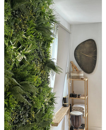 Une haie artificielle luxuriante Fraîcheur de jardin vertical UV 100x100 cm recouvre un mur d'une pièce lumineuse. A proximité, une horloge murale triangulaire moderne est suspendue au-dessus d'une étagère en bois avec des serviettes, un djembé, des paniers et de petites pièces décoratives.