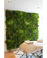 Une table de massage avec des serviettes pliées se dresse devant un jardin vertical UV Kunsthaag Freshness 100x100 cm dans une pièce lumineuse et moderne avec des murs blancs et un tapis géométrique.