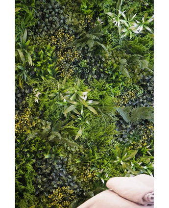 Une toile de fond luxuriante et texturée est créée par le mur de jardin vertical UV Kunsthaag Freshness 100x100 cm, densément recouvert de feuillage vert. Deux serviettes beiges pliées sont visibles en bas à droite.