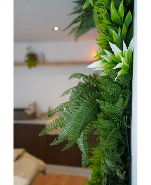 Un gros plan de plantes artificielles UV Fraîcheur de haie artificielle verte luxuriante de 100 x 100 cm orne un mur intérieur vertical, avec un arrière-plan flou montrant des meubles neutres et une petite plante suspendue.