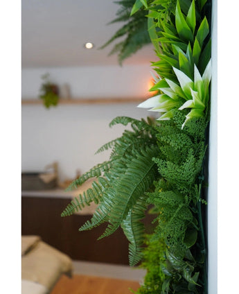 Un gros plan de plantes artificielles UV Fraîcheur de haie artificielle verte luxuriante de 100 x 100 cm orne un mur intérieur vertical, avec un arrière-plan flou montrant des meubles neutres et une petite plante suspendue.