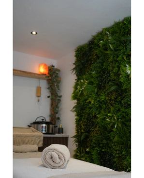 Un espace spa tranquille avec une table de massage, un éclairage apaisant, une lampe au sel de l'Himalaya sur une étagère et un mur végétal vert UV 100x100 cm Fraîcheur de Haie Artificielle luxuriante pour plus de sérénité.