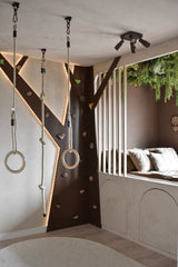 Une salle de jeux moderne pour enfants comprend un mur d'escalade, des anneaux de gymnastique, une corde et un coin lecture confortable avec des coussins et des lattes verticales. La haie artificielle Lush Deco Deluxe 100x100 cm UV ajoute de la verdure au plafond et complète les tons neutres et l'éclairage ludique de la pièce.