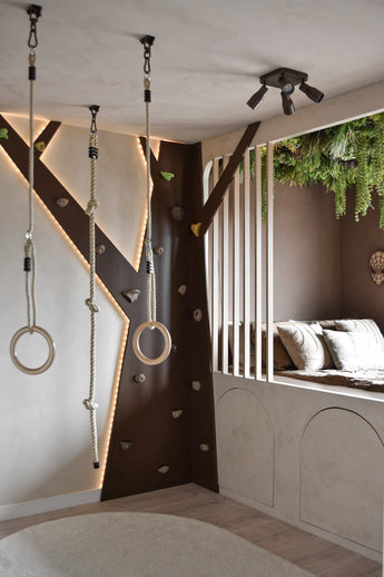 Une salle de jeux moderne pour enfants comprend un mur d'escalade, des anneaux de gymnastique, une corde et un coin lecture confortable avec des coussins et des lattes verticales. La haie artificielle Lush Deco Deluxe 100x100 cm UV ajoute de la verdure au plafond et complète les tons neutres et l'éclairage ludique de la pièce.