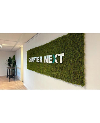 Un accent intérieur luxueux : un présentoir mural vert avec des cadres UV Kunsthaag Nordic Moss 50x50 cm, avec « CHAPTER NEXT » en lettres 3D grasses. Une table décorée de plantes complète la scène raffinée.