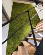 Un escalier moderne avec des marches en bois et des balustrades noires se dresse à côté d'un mur végétal en Kunsthaag Nordic Mos 50x50 cm UV et ajoute une touche naturelle et structurée à l'intérieur luxueux.