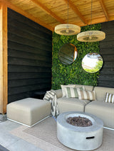 Coin salon extérieur moderne avec un canapé beige, des coussins rayés et une table ronde avec foyer. Deux miroirs ronds sont accrochés à une haie artificielle Volta, mur végétal UV 100x100 cm sous un plafond en bois et lampes suspendues en osier.