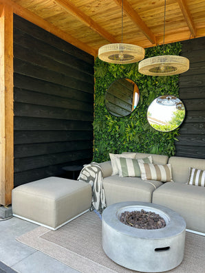 Coin salon extérieur moderne avec un canapé beige, des coussins rayés et une table ronde avec foyer. Deux miroirs ronds sont accrochés à une haie artificielle Volta, mur végétal UV 100x100 cm sous un plafond en bois et lampes suspendues en osier.