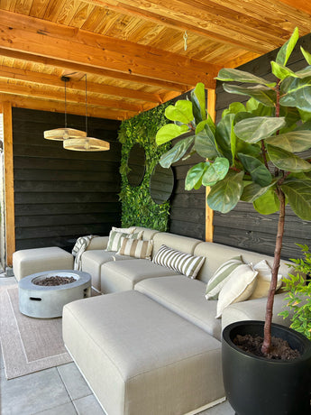 Un coin salon extérieur cosy avec un canapé beige, des coussins rayés, un foyer rond, une grande plante en pot et une pergola en bois. Le mur est décoré de verdure avec l'Artificiel Haag Volta 100x100 cm UV pour plus de style.