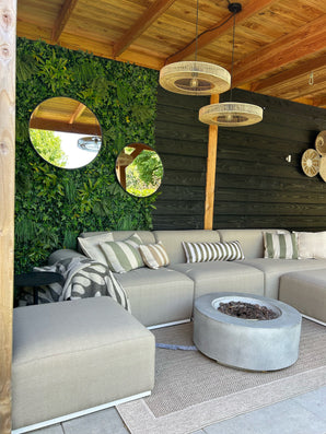 Une terrasse cosy avec un canapé beige, des coussins rayés, un foyer et des tapis tissés. Le vert UV de Kunsthaag Volta 100x100 cm est accroché au mur, des miroirs ronds et des lampes en osier sous une pergola en bois.