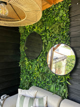 Un coin salon confortable avec un canapé beige et des coussins rayés se trouve sous un plafond en bois. Derrière, une haie artificielle de verdure luxuriante, un mur végétal UV Volta 100x100 cm avec deux miroirs ronds ajoute une touche naturelle au mur laqué noir.