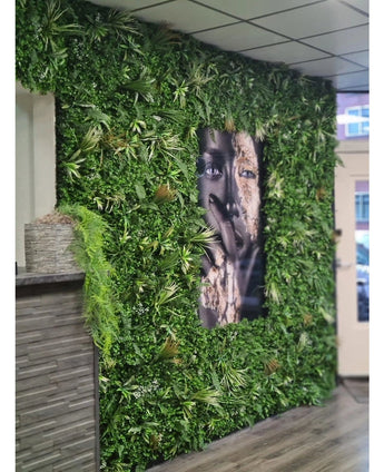 Un mur recouvert de tapis de haie artificielle Savanne 100x100 cm, avec de la verdure protégée contre les UV dans une jardinière en plastique Ø100, présente au milieu un grand portrait en noir et blanc. L'intérieur moderne présente un plafond carrelé et un comptoir en bois sur la gauche.