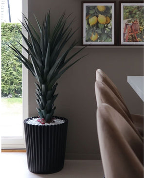 La plante artificielle Agave Pandora UV 120 cm, une grande plante verte épineuse dans un pot nervuré noir avec des pierres blanches, se trouve près de la fenêtre. Derrière, trois imprimés de fruits encadrés sont accrochés à une étagère murale au-dessus de chaises beiges et d'une table blanche.
