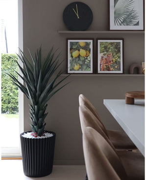 Un coin repas moderne avec des chaises beiges, une table blanche, une horloge murale et une étagère avec des photos encadrées de citrons et de feuilles. La grande plante artificielle Agave Pandora UV 120 cm ajoute de la verdure. La lumière naturelle entre par le côté gauche.