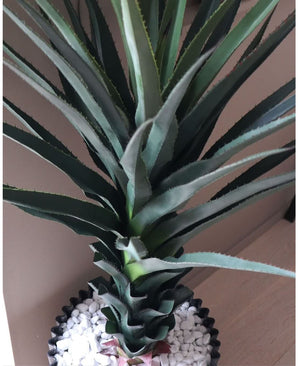 Un gros plan de la plante artificielle Agave Pandora UV 120 cm, avec des feuilles vertes épineuses dans un pot noir avec des galets blancs, exposée sur une surface claire contre un mur neutre.