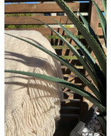 Un gros plan de la plante artificielle Agava Pandora UV 85 cm dans un pot avec des galets blancs se trouve à côté d'une couverture de couleur crème à franges drapée sur un sommier à lattes en bois, avec la lumière du soleil projetant des ombres.