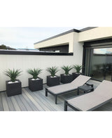 Un patio moderne comprend deux chaises longues grises et cinq jardinières carrées, chacune avec une plante artificielle Agava Pandora UV 85 cm, contre un mur lambrissé blanc et noir pour un look élégant et minimaliste.