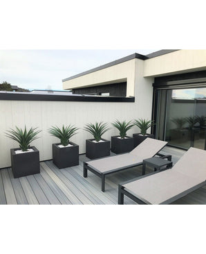 Un patio moderne comprend deux chaises longues grises et cinq jardinières carrées, chacune avec une plante artificielle Agava Pandora UV 85 cm, contre un mur lambrissé blanc et noir pour un look élégant et minimaliste.
