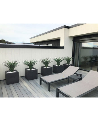 Un patio moderne comprend deux chaises longues grises et cinq jardinières carrées, chacune avec une plante artificielle Agava Pandora UV 85 cm, contre un mur lambrissé blanc et noir pour un look élégant et minimaliste.