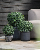 Trois buis artificiels verts ronds sur tige UV d30 cm dans des pots noirs nervurés se dressent sur une terrasse en bois à côté d'un canapé gris clair, avec un mur en bois sombre derrière.