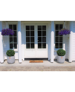 Les doubles portes blanches avec vitres sont flanquées de deux buis artificiels sur tige de style romain UV 65 cm dans de grands pots. Deux paniers suspendus avec des fleurs violettes et un paillasson complètent l'entrée.