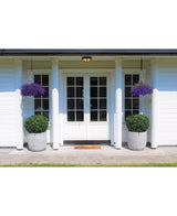 Des doubles portes blanches avec panneaux de verre ornent une maison blanche à quatre colonnes. Deux topiaires de Buis Artificiel sur tige UV d60 cm et deux paniers suspendus à fleurs violettes décorent l'entrée, avec un paillasson devant les portes.