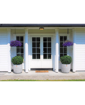 Des doubles portes blanches avec panneaux de verre ornent une maison blanche à quatre colonnes. Deux topiaires de Buis Artificiel sur tige UV d60 cm et deux paniers suspendus à fleurs violettes décorent l'entrée, avec un paillasson devant les portes.