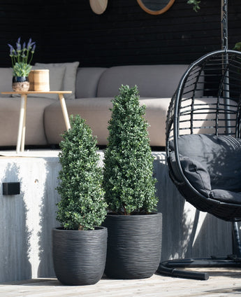 Deux arbres artificiels en forme de pyramide de buis UV de 90 cm se trouvent sur une terrasse près d'un banc extérieur et d'une chaise suspendue ronde en osier avec un coussin noir, tandis qu'une petite table avec une plante est en arrière-plan.
