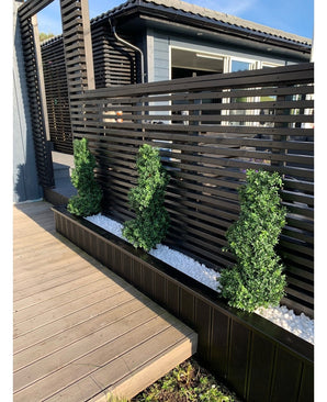 Trois arbustes topiaires artificiels de style romain en spirale de buis UV de 90 cm se dressent dans un lit étroit de galets blancs, contre une clôture d'intimité moderne à lattes noires à côté d'un patio en bois et d'une maison contemporaine.