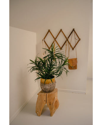 La Plante Artificielle Dracaena Reflexa en pot UV 63 cm décore une pièce minimaliste aux murs blancs, posée sur un tabouret en bois naturel. Une étagère murale en bois à l'arrière accueille un petit sac marron et des tissus crème pour un intérieur tropical.