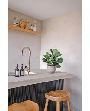 Un plan de travail moderne avec un robinet en laiton, deux distributeurs de savon, la plante en pot Artificielle Feuille de Tabac UV 45 cm et deux tabourets en bois. Une étagère flottante avec des pots et des bouteilles est montée sur un mur de couleur claire.