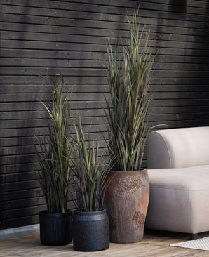 Trois plantes artificielles de gazon artificiel Bordeaux UV de 90 cm avec de longues feuilles vertes minces se tiennent à côté d'un canapé beige sur une terrasse en bois, contre un mur de lattes de bois sombre.