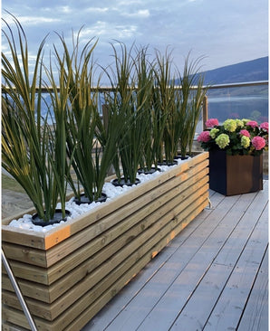 Une jardinière en bois avec gazon artificiel vert UV 120 cm et pierres blanches se trouve sur un balcon avec balustrade en verre ; à proximité, un pot carré noir d'hortensias en fleurs roses, verts et blancs surplombe la vue pittoresque.