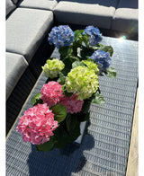 La plante Hortensia Artificielle Vert UV 35 cm se dresse sur une table en osier avec un plateau en verre à côté d'un canapé d'extérieur gris, avec des hortensias verts réalistes qui s'illuminent au soleil.