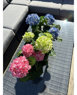 La plante Hortensia Artificielle Vert UV 35 cm se dresse sur une table en osier avec un plateau en verre à côté d'un canapé d'extérieur gris, avec des hortensias verts réalistes qui s'illuminent au soleil.