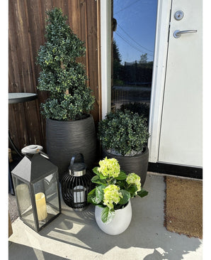Une plante d'hortensia artificiel vert UV de 35 cm dans un pot blanc avec des fleurs jaune-vert se dresse sur la véranda près d'une porte marron, à côté de deux plantes dans un pot noir et de deux lanternes décoratives avec des bougies.