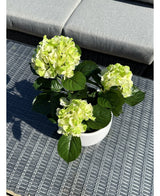 L'hortensia artificiel vert UV 35 cm, avec des fleurs vert clair et des feuilles vert foncé, se dresse sur une table d'extérieur grise tressée avec un banc rembourré en arrière-plan.