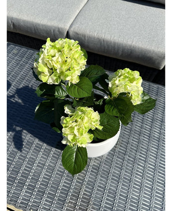 L'hortensia artificiel vert UV 35 cm, avec des fleurs vert clair et des feuilles vert foncé, se dresse sur une table d'extérieur grise tressée avec un banc rembourré en arrière-plan.