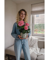 Une femme aux cheveux blonds bouclés, vêtue d'une veste en jean et d'un jean, se tient à l'intérieur et sourit tout en tenant la plante d'hortensia artificielle rose UV de 35 cm. Au fond il y a un canapé et une fenêtre.