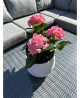 La Plante Artificielle Hortensia Rose UV 35 cm, un pot blanc avec des fleurs d'hortensia rose vif, se dresse sur une table d'extérieur grise tressée avec un canapé gris avec des coussins en arrière-plan, éclairé par la lumière du soleil et des ombres douces.