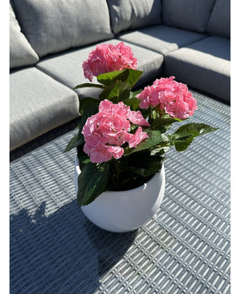La Plante Artificielle Hortensia Rose UV 35 cm, un pot blanc avec des fleurs d'hortensia rose vif, se dresse sur une table d'extérieur grise tressée avec un canapé gris avec des coussins en arrière-plan, éclairé par la lumière du soleil et des ombres douces.